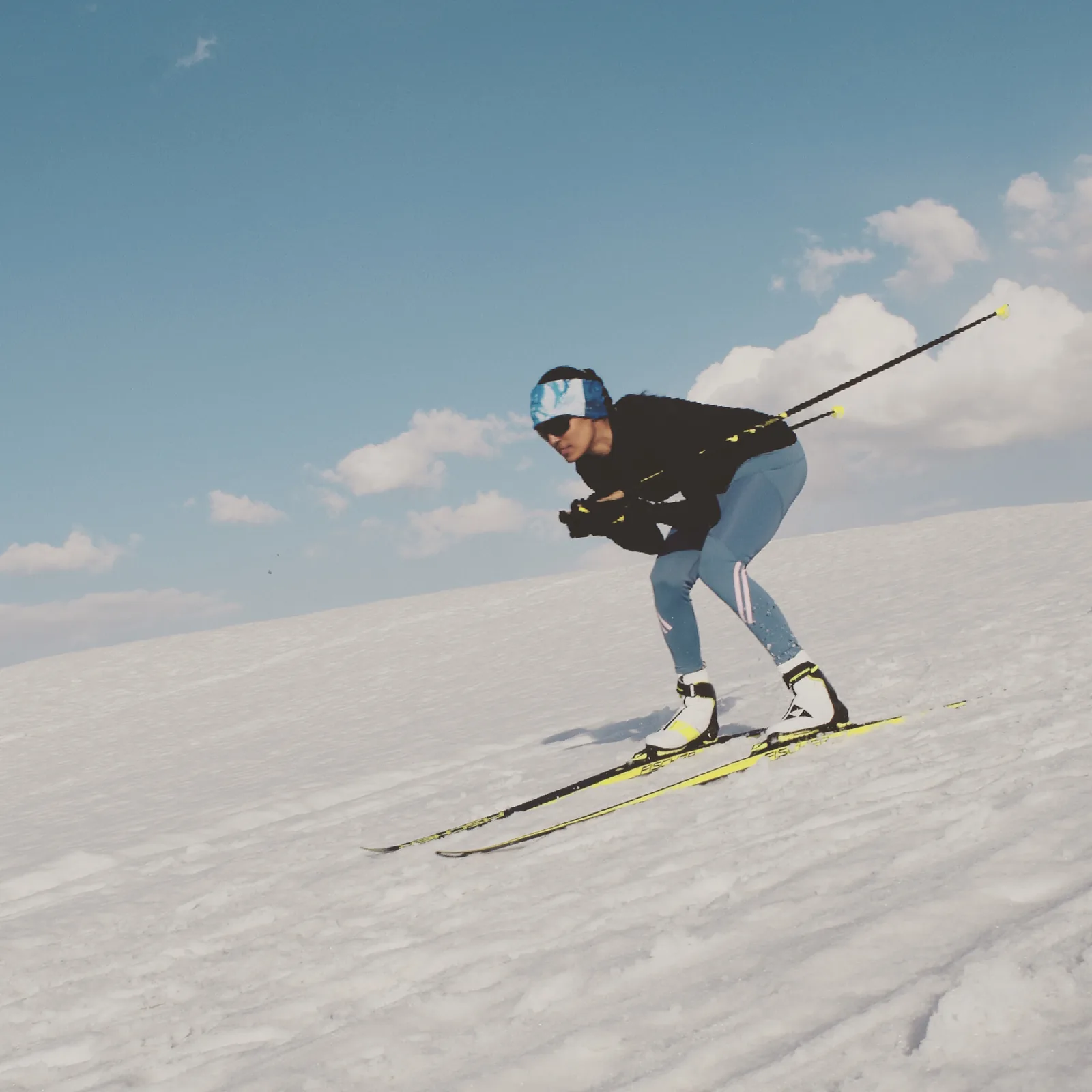 Bhavani Thekkada low in a tuck on skis, carrying speed across an open slope against a clear sky.