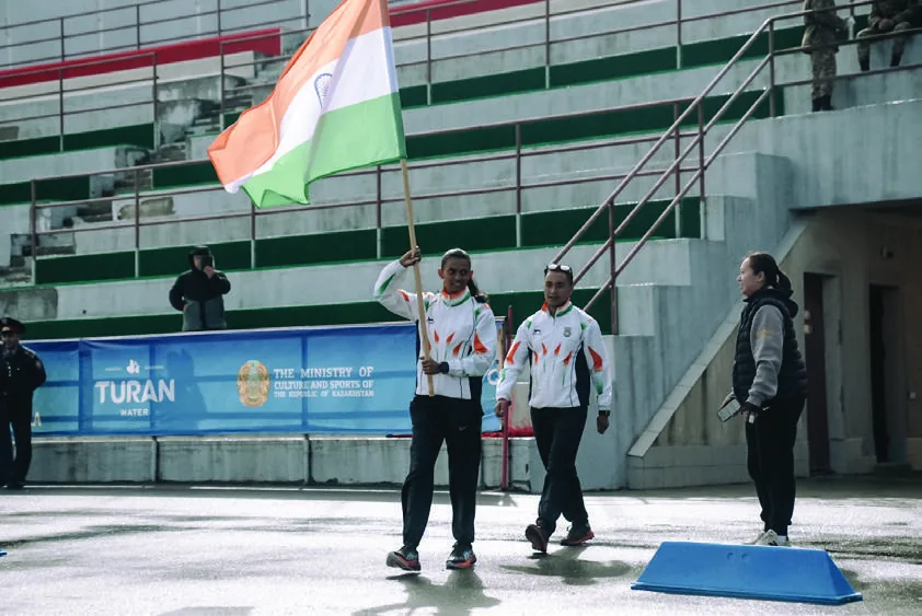 Athletes in white India kit on an outdoor rink, one holding the Indian flag open above a stadium barrier.