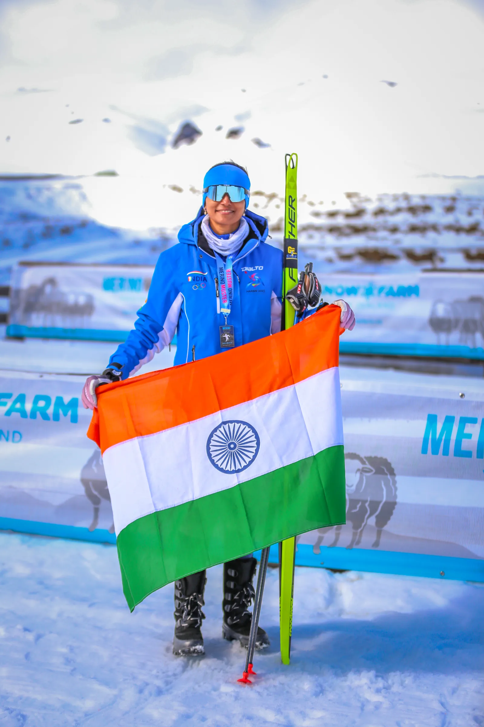 Bhavani Thekkada holding the Indian flag open in front of the Snow Farm and Merino Muster banners, her skis beside her.
