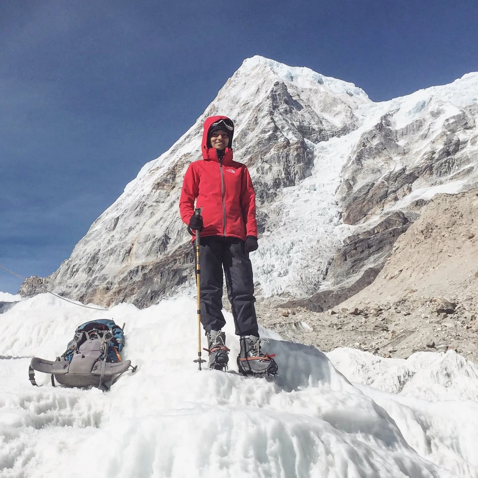 Bhavani Thekkada in a red down jacket and crampons standing on a glacier below a steep face of snow and rock.