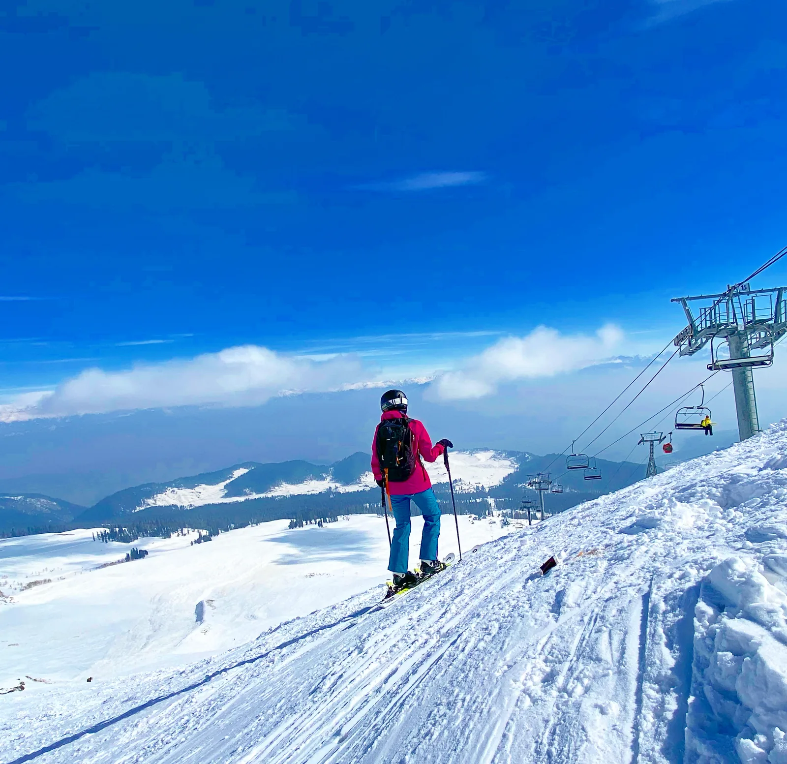 A skier in a pink jacket at the top of a groomed piste above a wide snowfield, a chairlift running down the slope beside her.