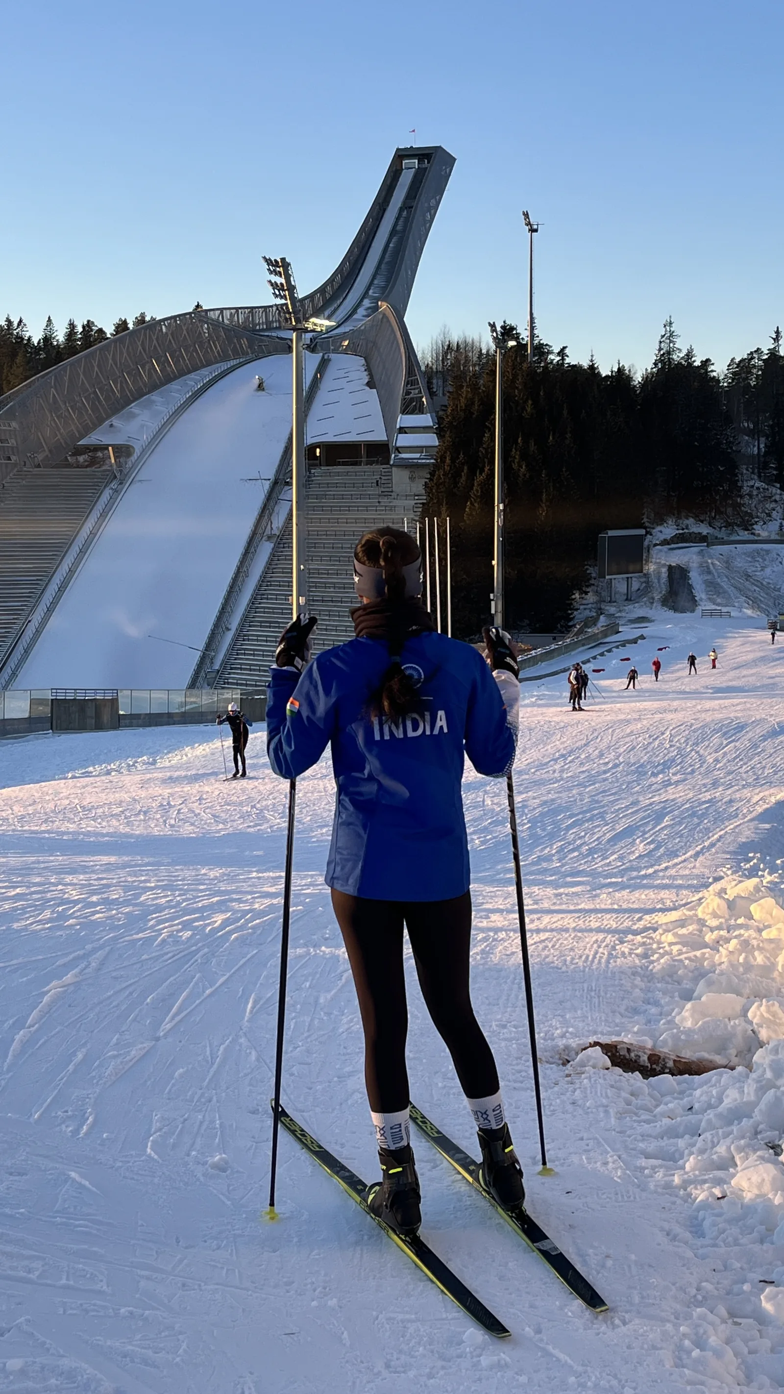 Bhavani Thekkada standing on her skis in an India training top below a large ski jump tower.