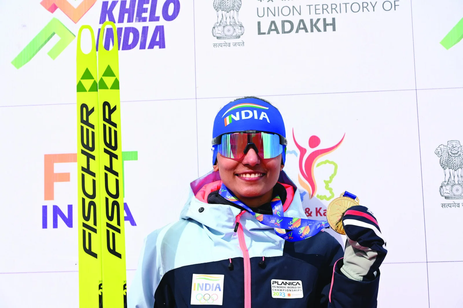 Bhavani in team kit and mirrored glasses in front of a Khelo India Ladakh backdrop, upright skis flanking her.