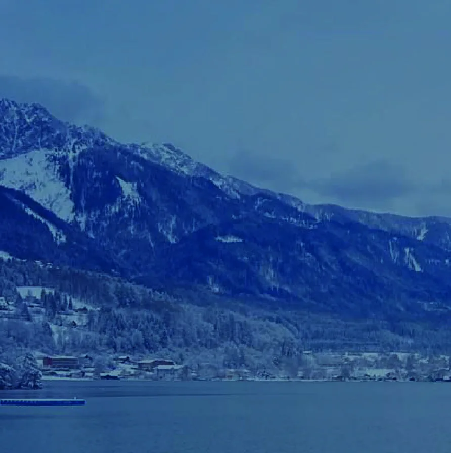 A still alpine lake below a long snow-streaked mountain ridge in flat winter light.