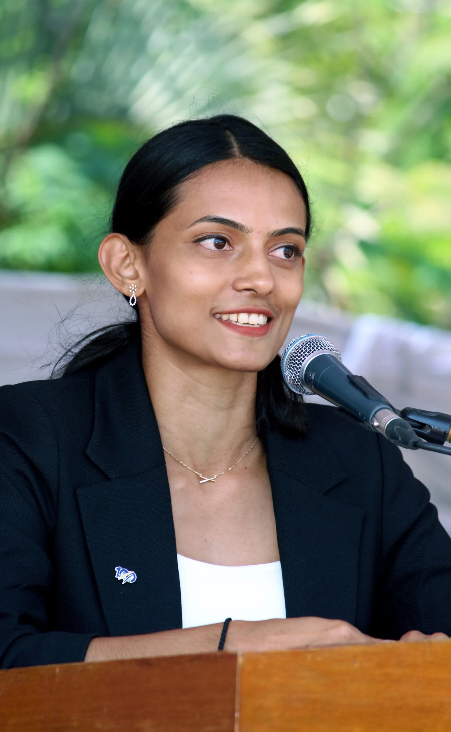 Bhavani Thekkada speaking at a lectern with a microphone in front of her, in a dark blazer, greenery behind.