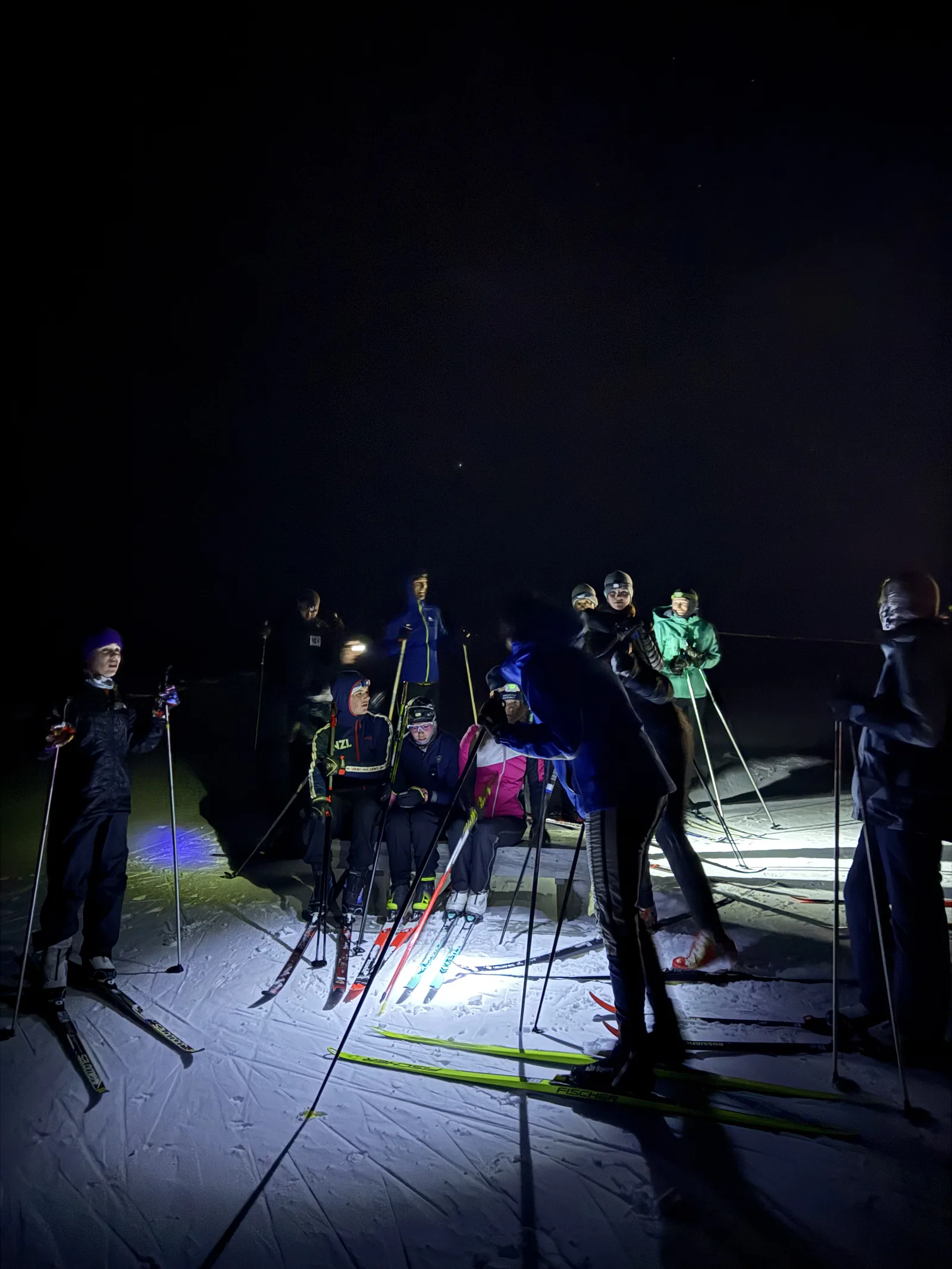 Skiers training on a floodlit trail after dark, headlamps and lit snow against a black sky.