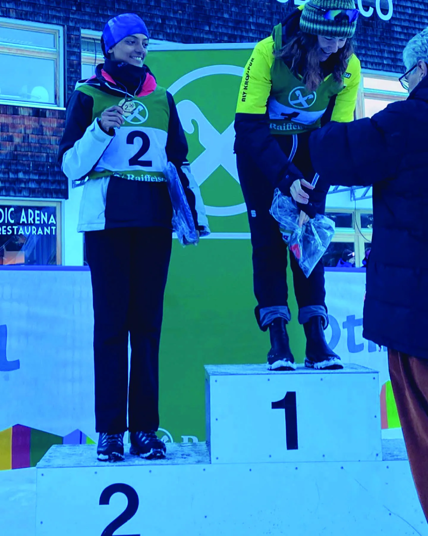Two athletes on a numbered race podium, one being handed a prize at the top step.