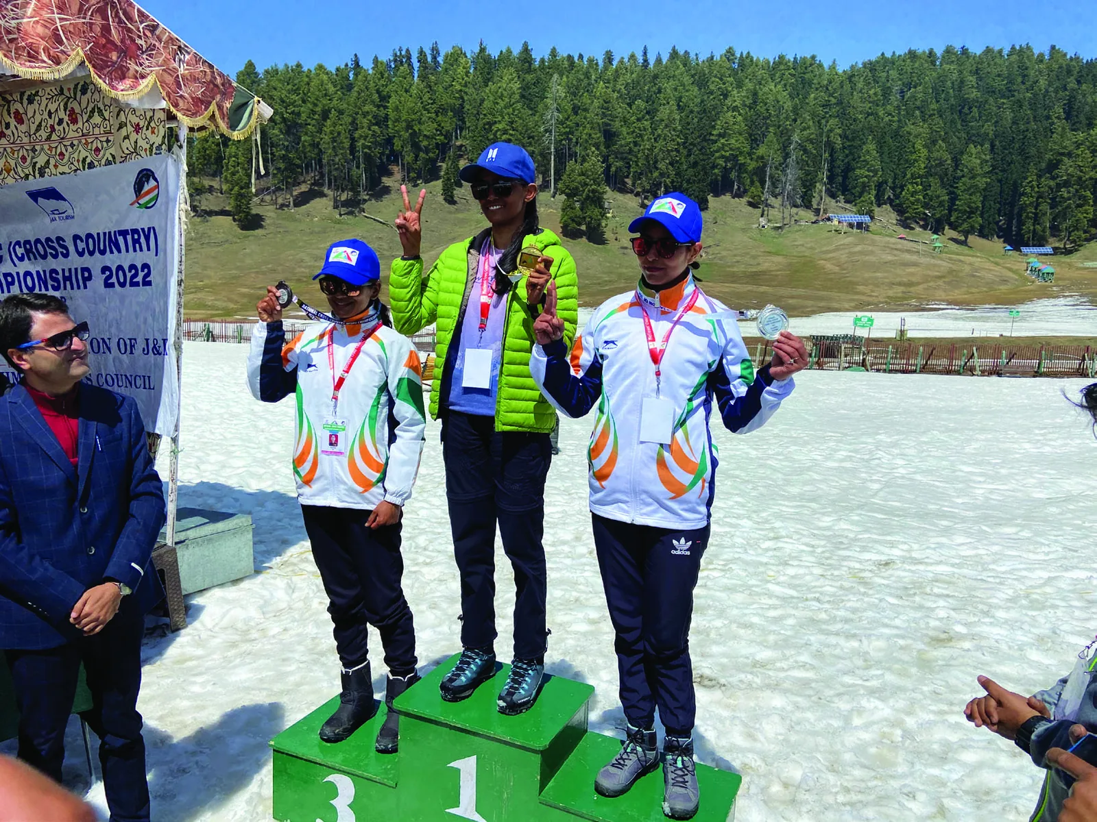 Three medallists on a three-step podium on snow in front of a Cross Country Championship 2022 banner, mountains and pine forest behind.