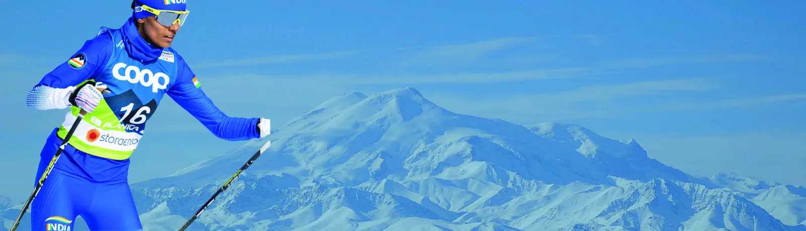Bhavani racing in a blue World Championships bib, arm extended mid-stride, against a wide snow-covered mountain massif.