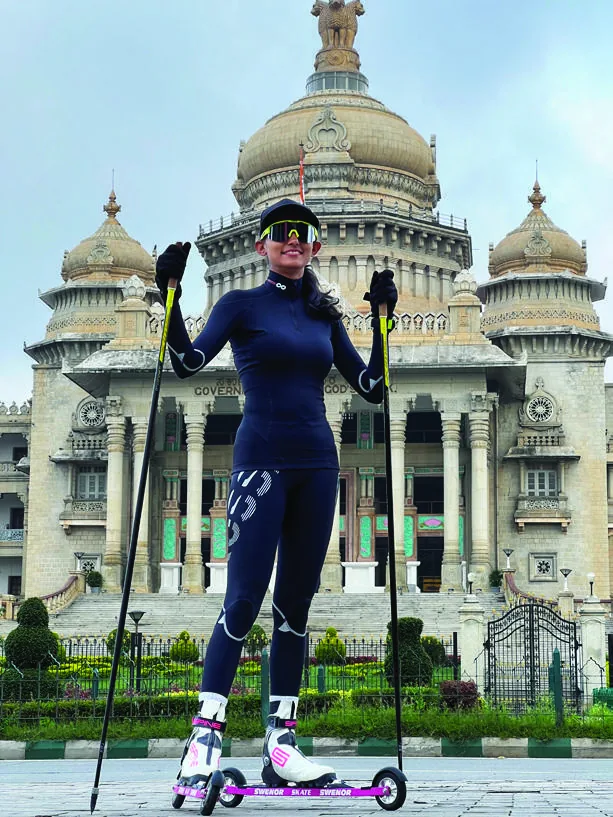 Bhavani on roller skis with poles raised, on the road in front of the domed Vidhana Soudha in Bengaluru.