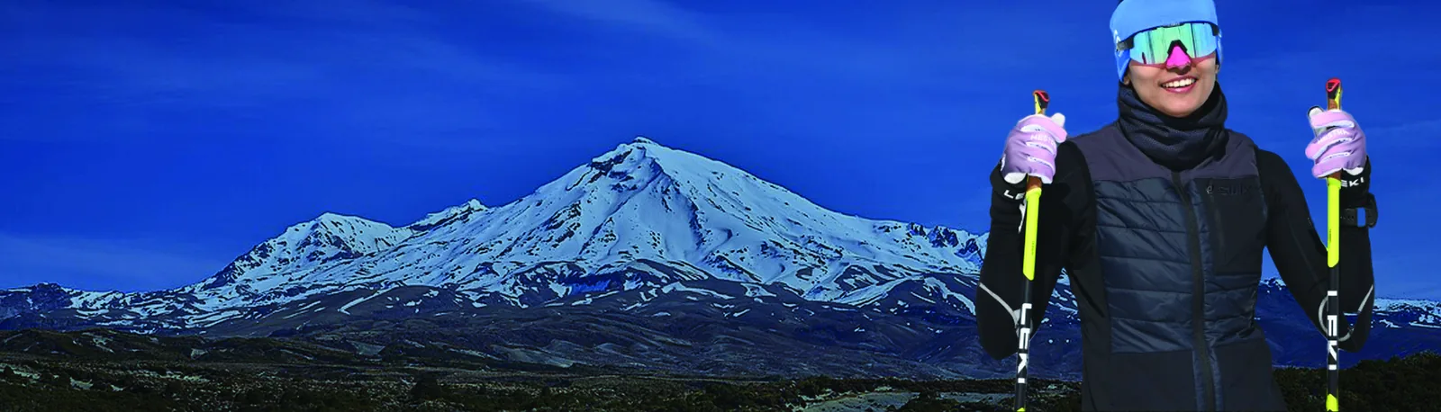 Mount Ruapehu under a deep blue sky, its snow cap above dark volcanic ground, with Bhavani standing at the right holding ski poles.