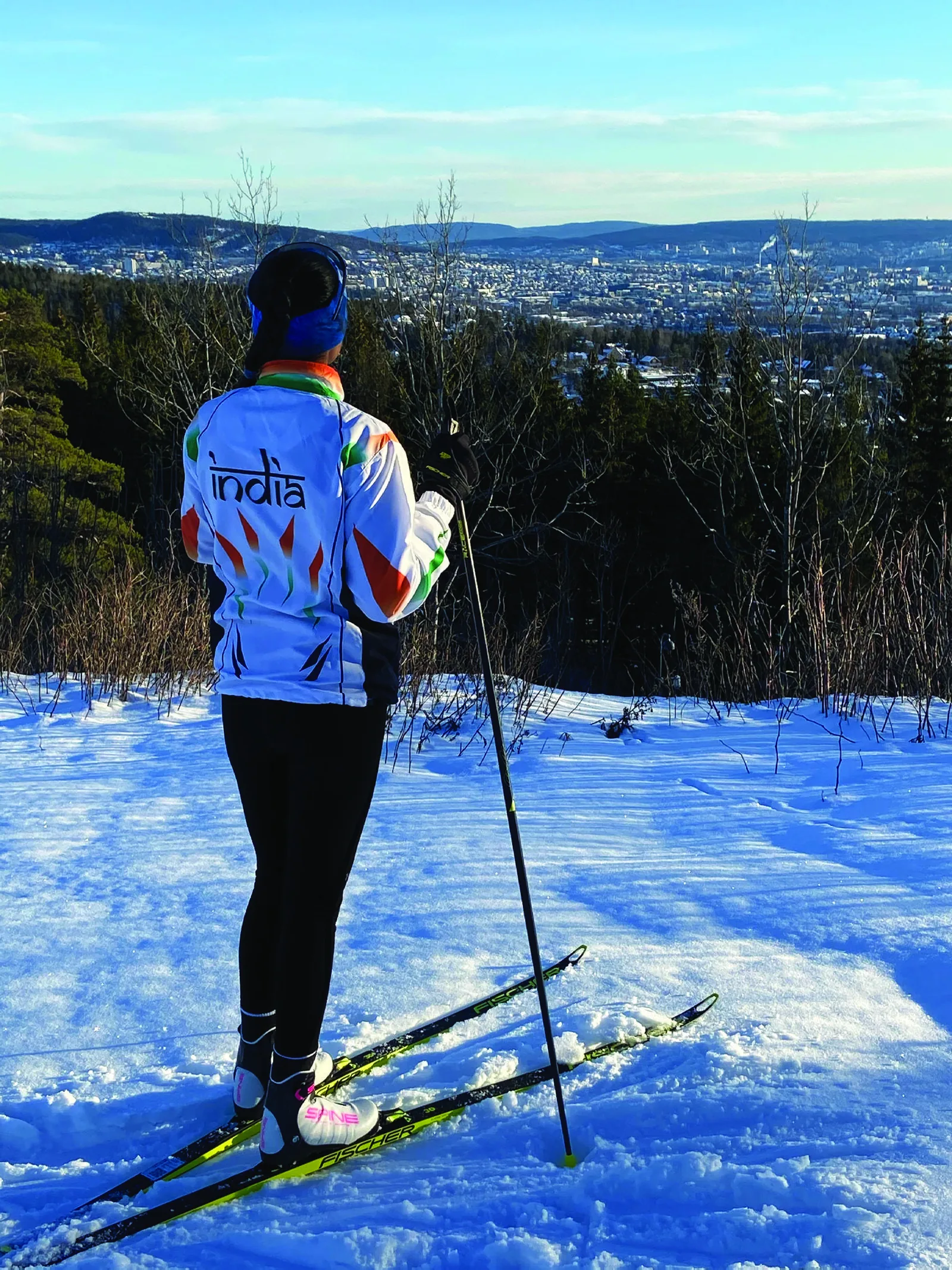 Bhavani seen from behind in an India team jacket, standing on skis at the top of a slope with a city spread across the valley below.