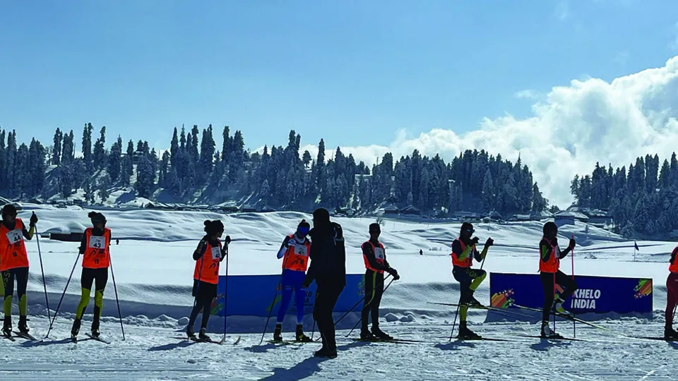 A line of skiers in orange bibs waiting at a race start on open snow, forest and mountains behind.