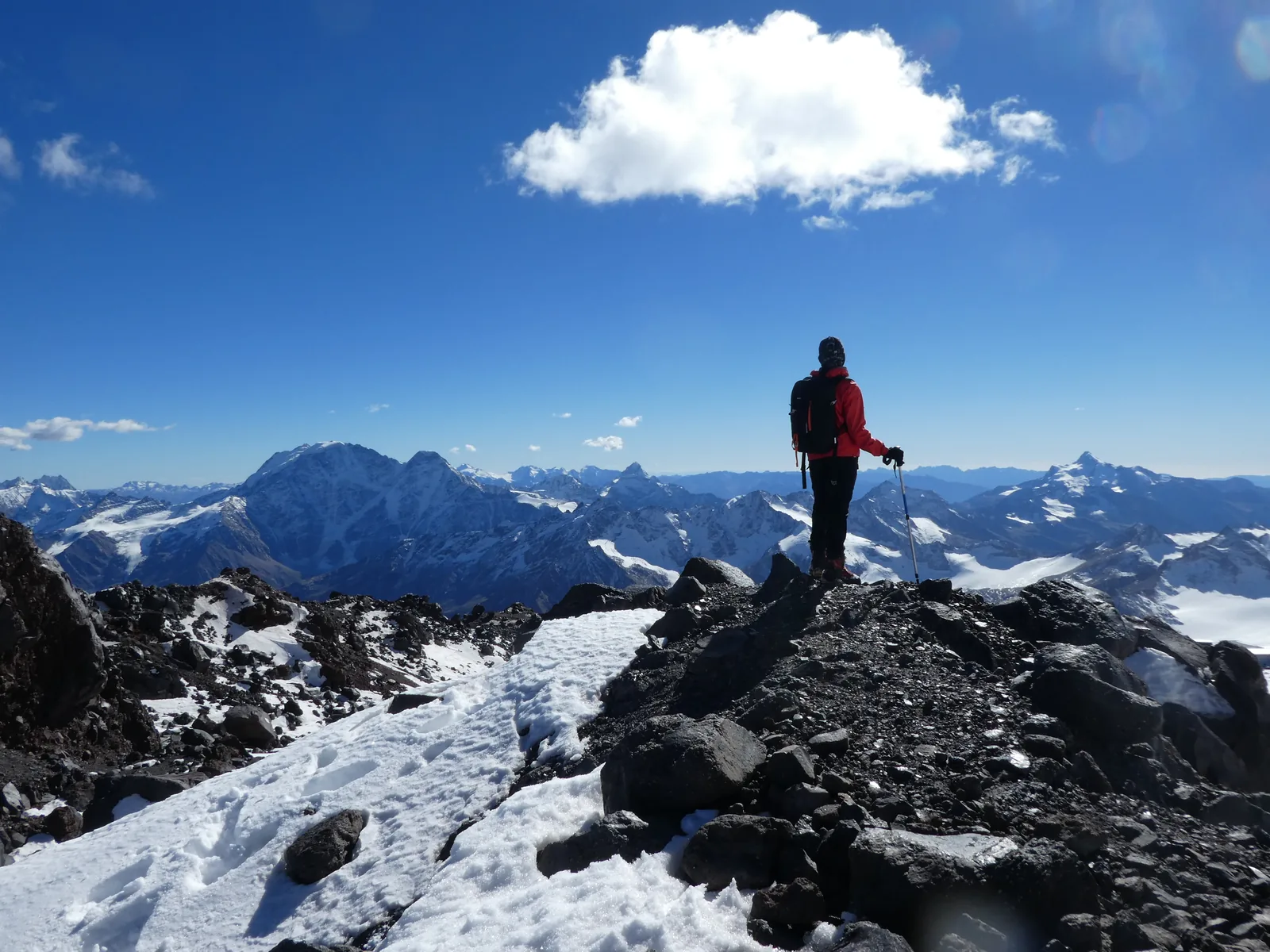 A single climber standing on a rocky summit ridge with a horizon of snow peaks behind and cloud above.