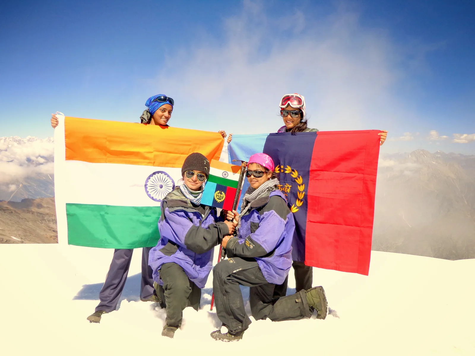 Four climbers on a snow summit holding the Indian tricolour and an NCC flag open between them, a ridge line and cloud below.