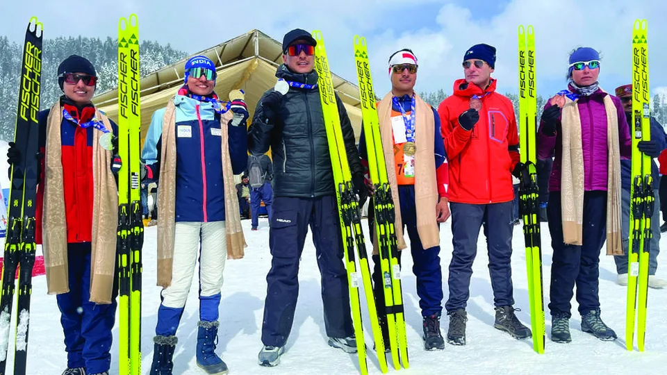 A group of skiers and coaches standing shoulder to shoulder holding their skis upright, wrapped in shawls against the cold.